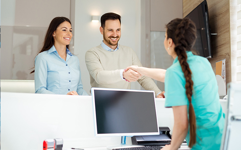 Couple Smiling At Dental Reception Desk While Man Shakes Hands With Staff Member In Modern Office