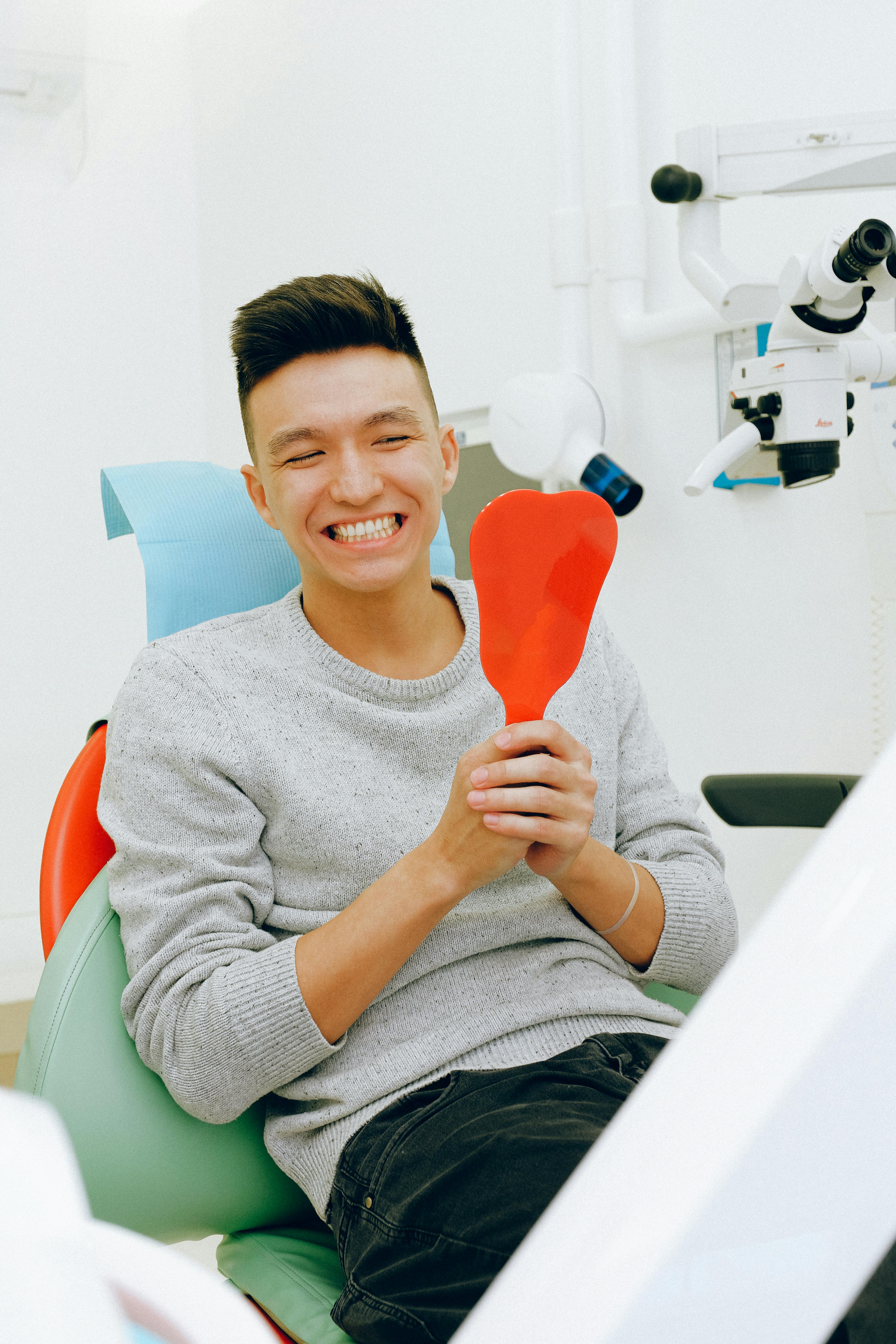 Photo Young Man Smiling In Dental Chair Holding Red Hand Mirror What Is A Smile Makeover Consultation With Dental Equipment In Background
