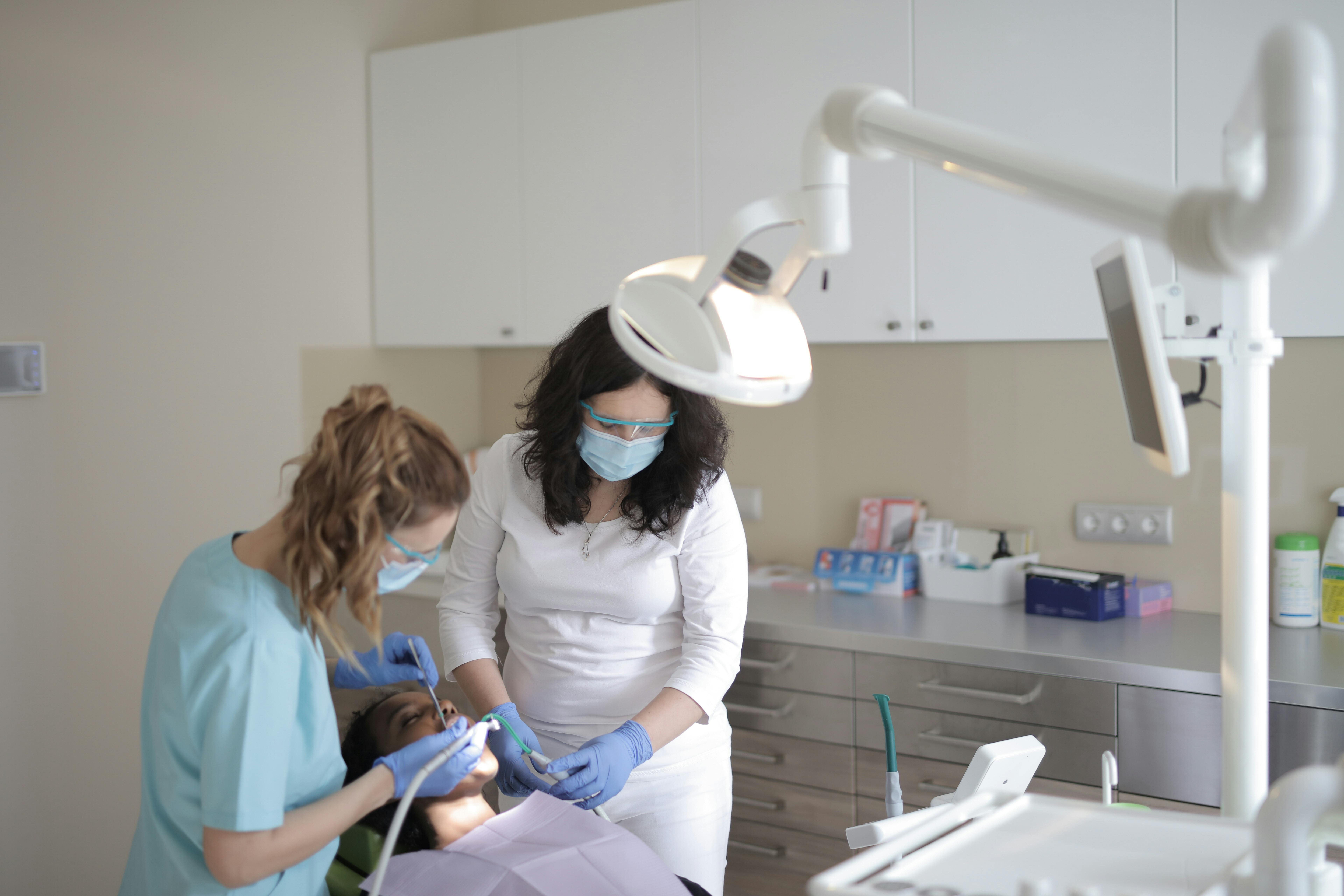 Photo Two Dentists Wearing Masks And Gloves Treating Patient In Chair Under Exam Light Demonstrating Dental Emergency Tips In Modern Clinic