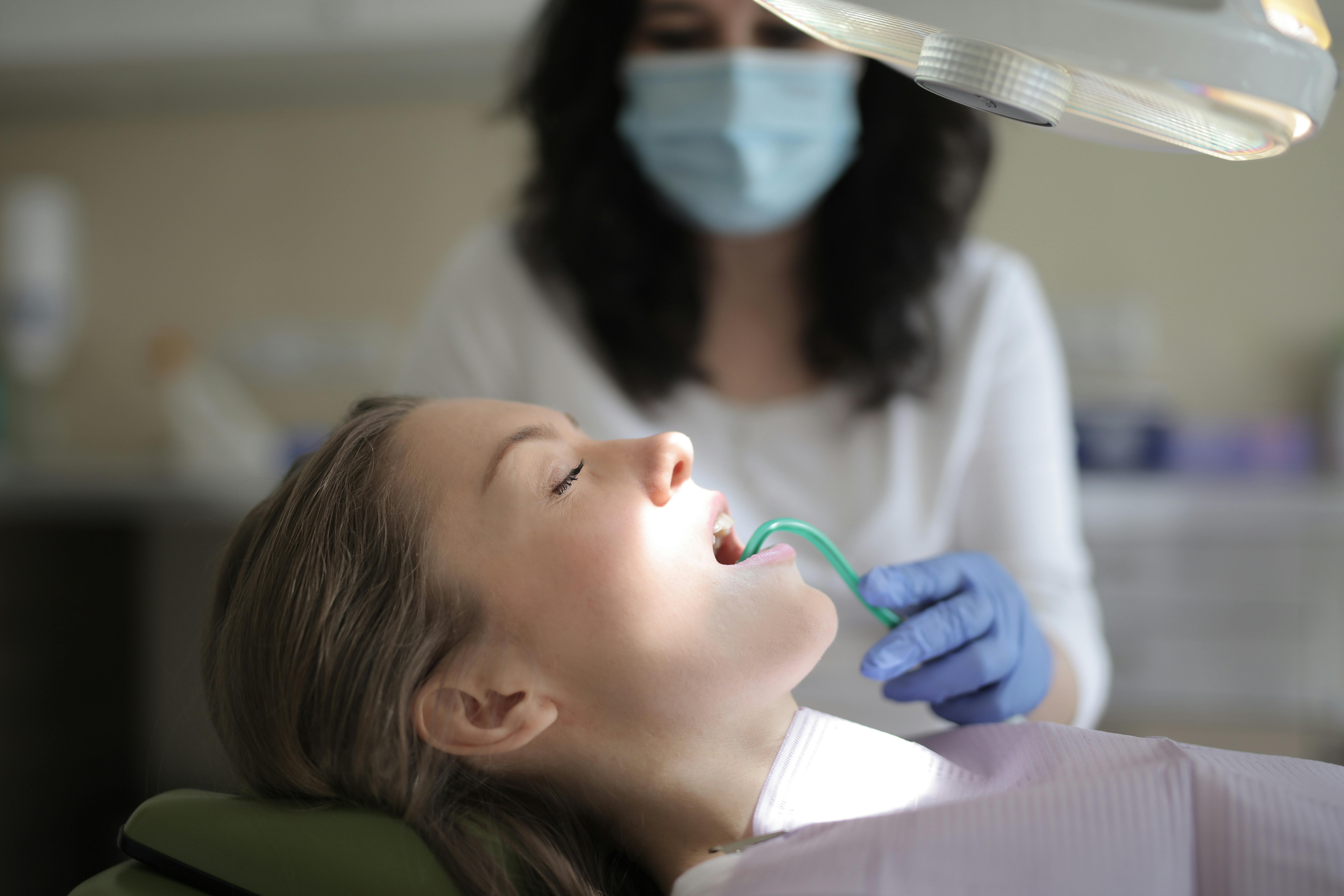Photo Dentist Wearing Mask And Gloves Examining Patient Mouth With Suction Tool Under Overhead Light