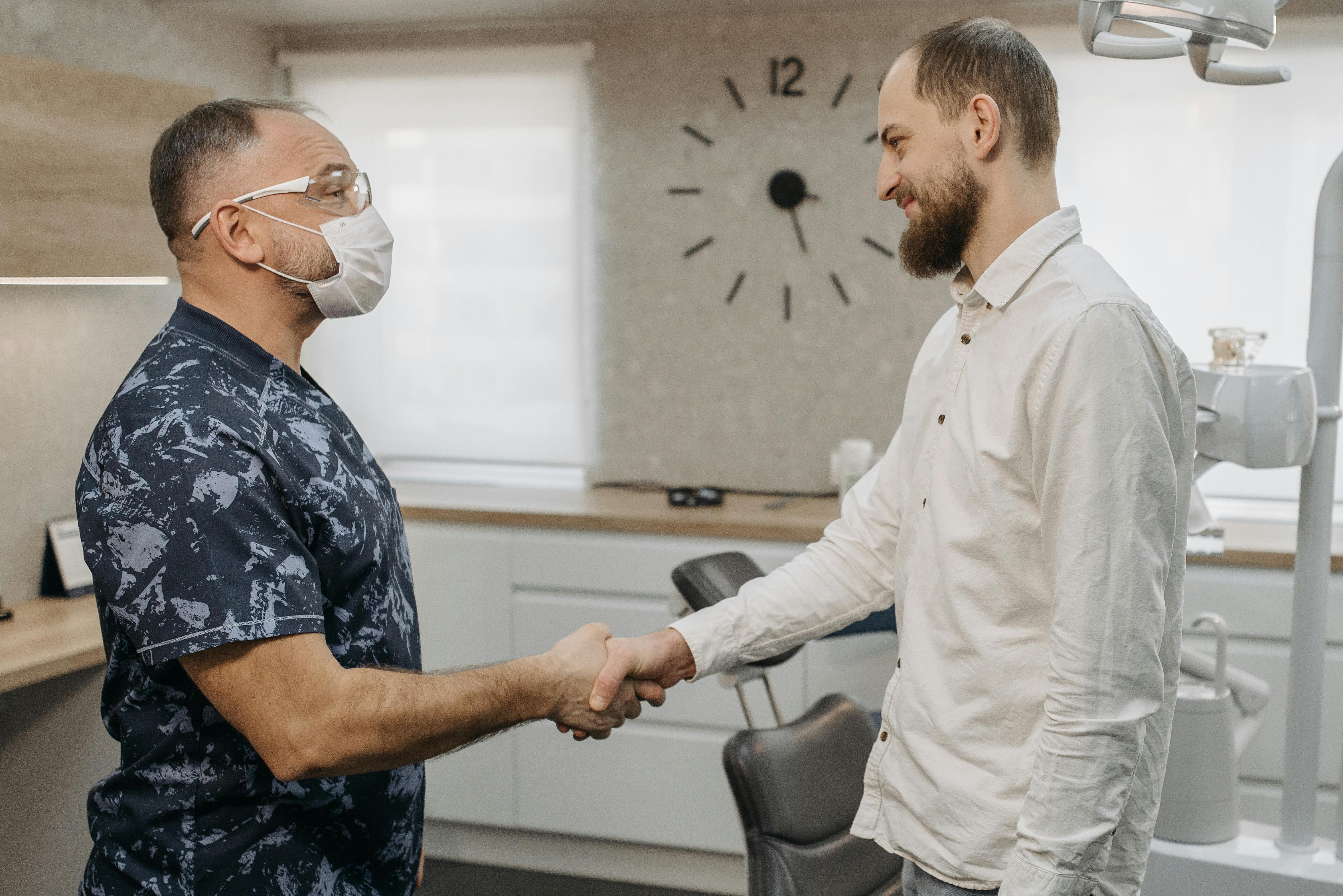 Photo Of Dentist Wearing Mask Shaking Hands With Man In Dental Office With Chair And Wall Clock