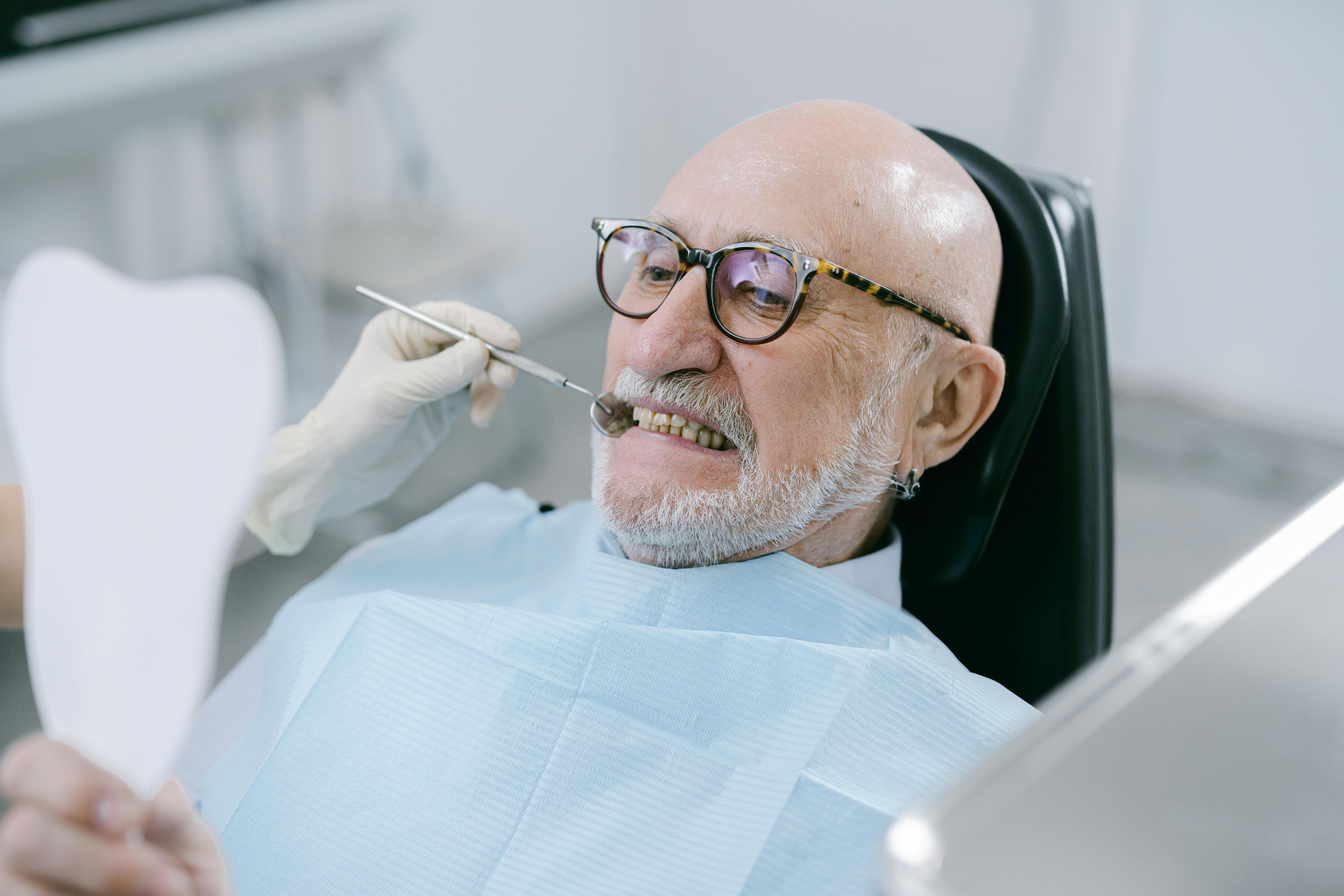 Photo Of Elderly Man In Dental Chair Holding Mirror While Dentist Examines Teeth With Dental Tool