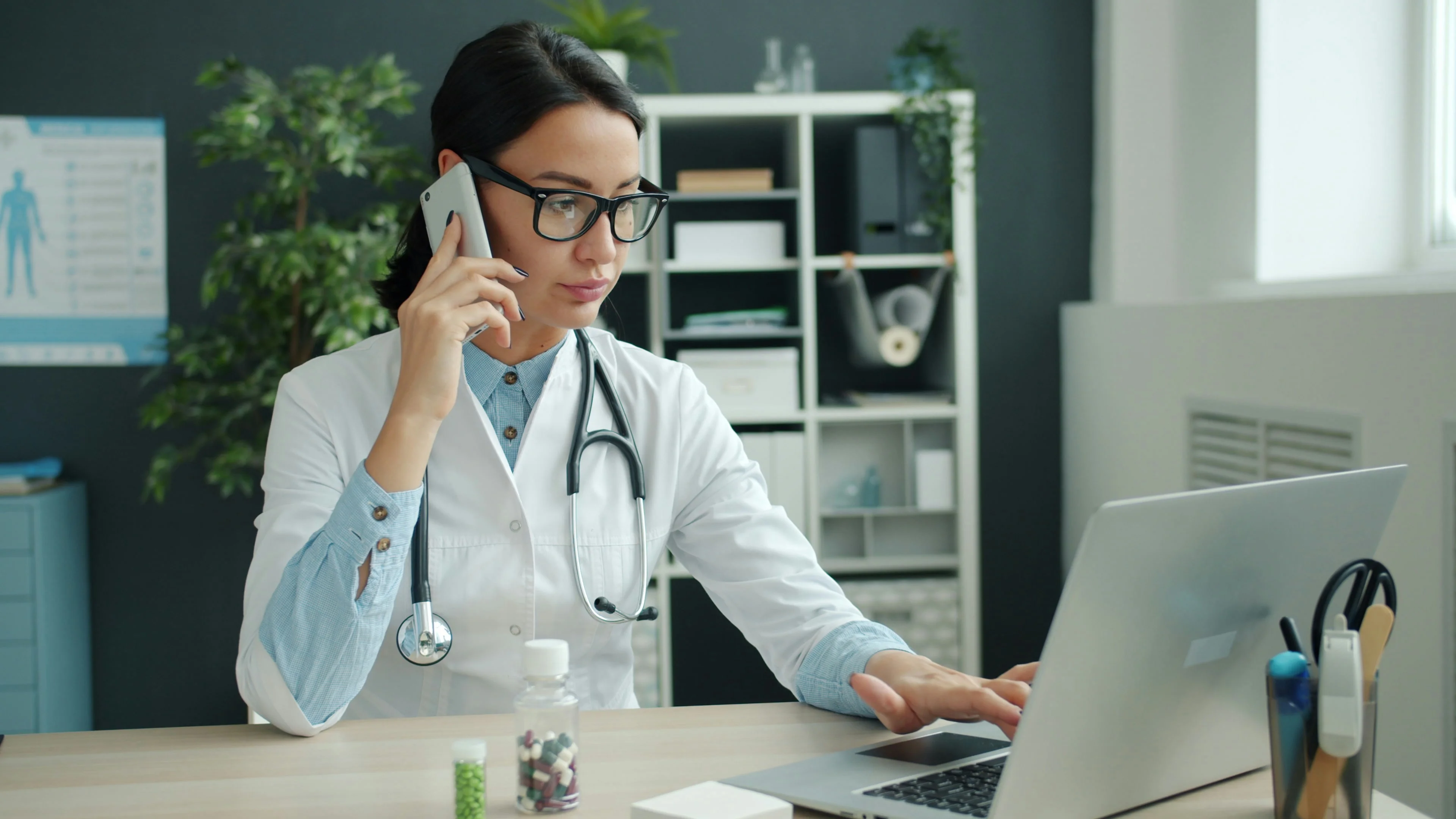 Photo Doctor Talking On Phone While Using Laptop At Desk With Medical Supplies And Stethoscope In Office