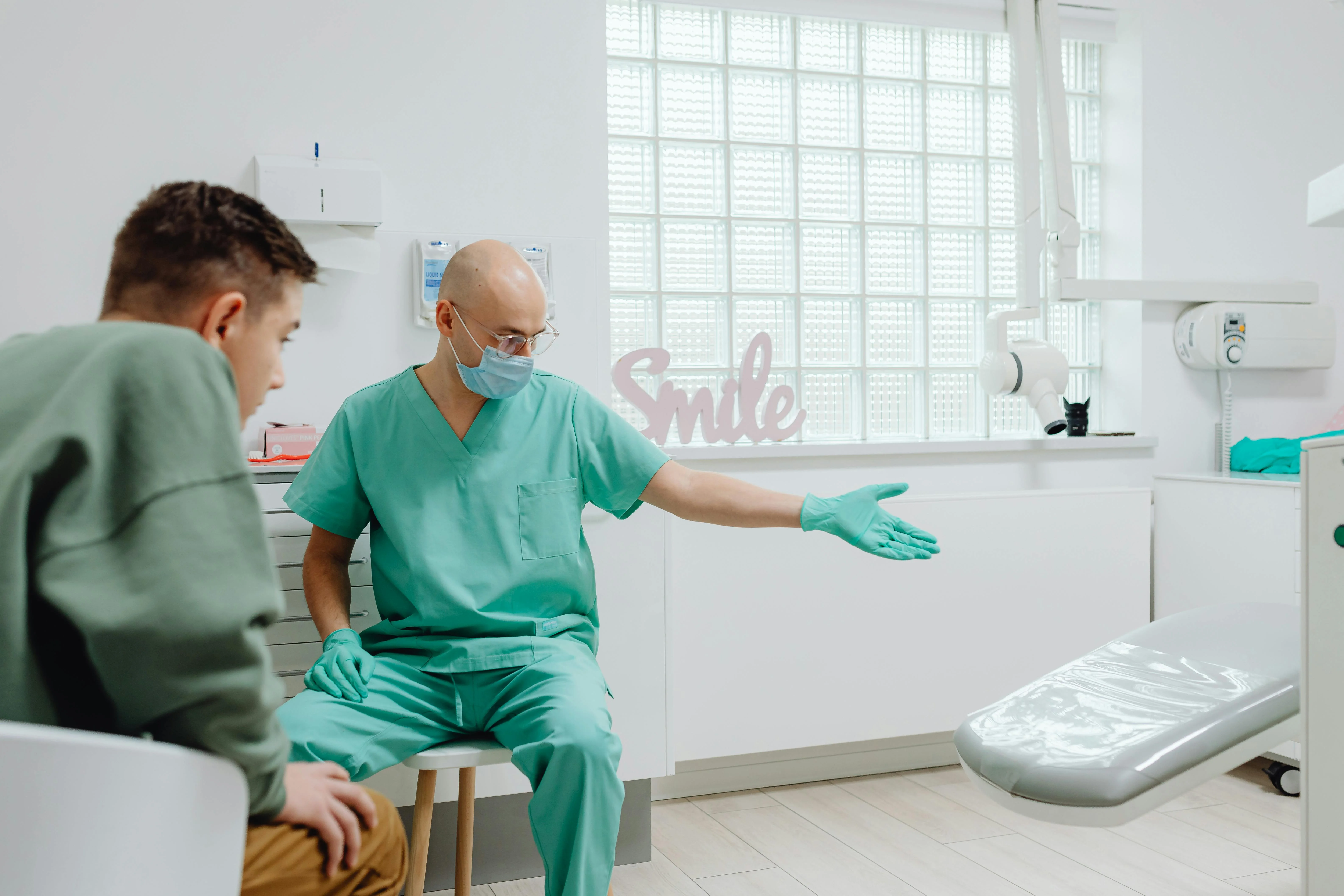 Photo Dentist Gesturing Toward Dental Chair While Patient Sits Nearby Showing How To Handle Dental Emergency Anxiety In A Bright Clinic