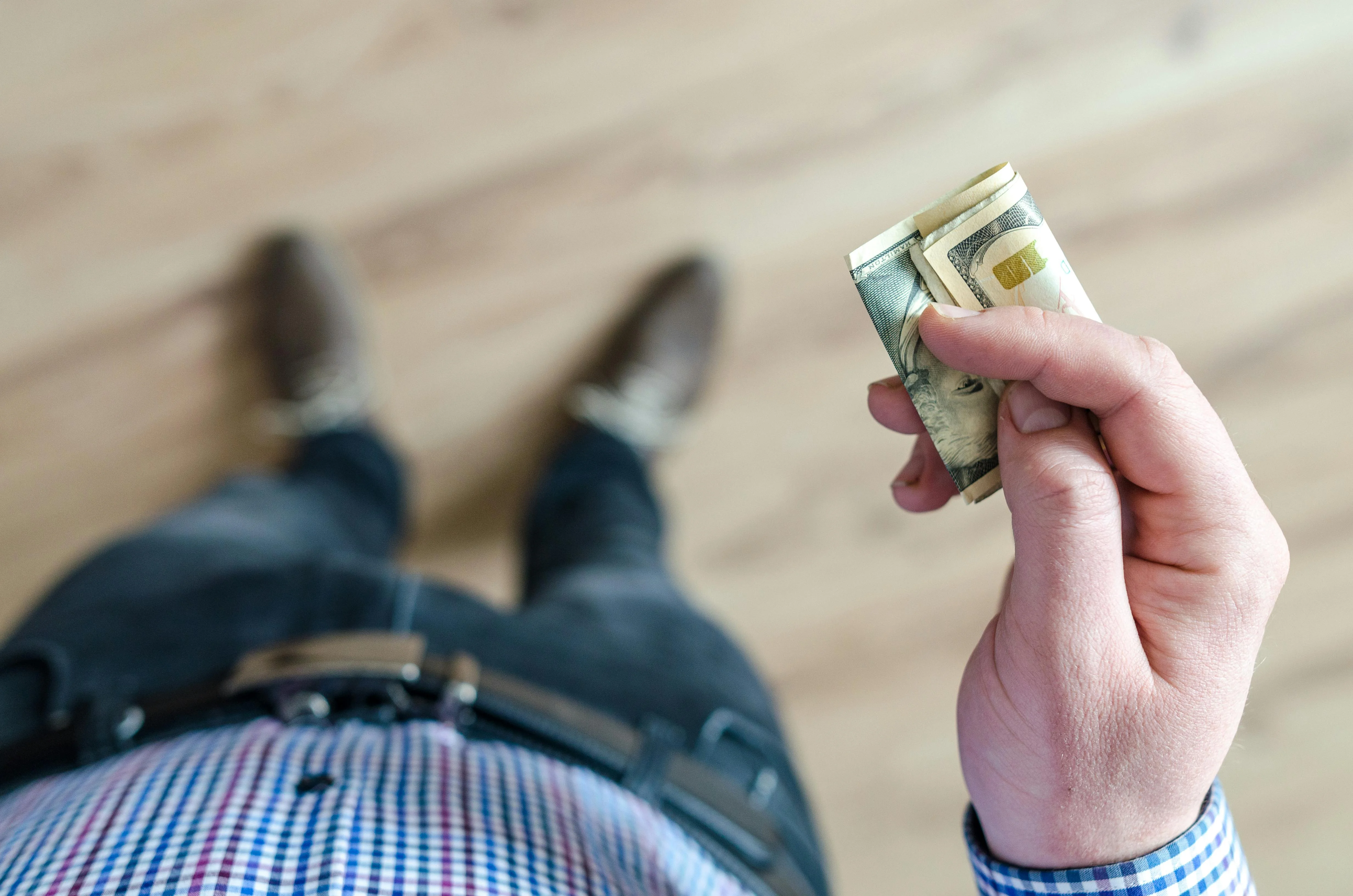Photo Man Holding Folded Cash While Standing On Wood Floor Wearing Blue Checkered Shirt And Dark Pants