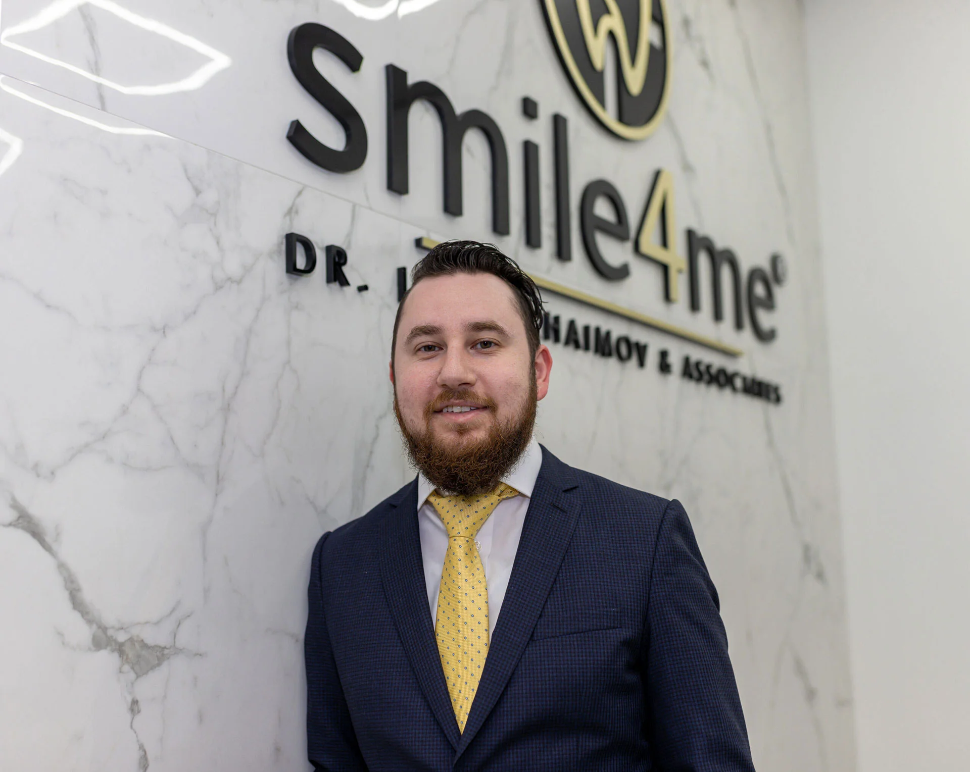 Man In Navy Suit And Yellow Tie Standing In Front Of Smile4me Dental Care Logo On Marble Wall Background Looking At Camera