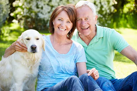 Smiling Middle Aged Couple Sitting On Grass Outdoors With Golden Retriever Dog Enjoying Sunny Day And Looking Happy Together
