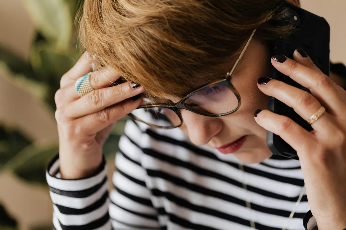 Woman Wearing Glasses And Striped Shirt Talking On Phone Looking Concerned While Asking Do Dental Implants Fall Out