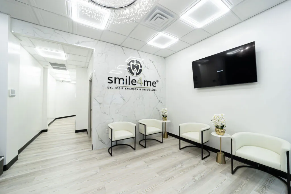 Modern Dental Office Waiting Room With White Chairs, Flat Screen TV, Marble Wall, And Smile4Me Logo Displayed Behind Reception Area.